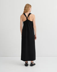 A woman viewed from the back wearing a sleeveless black maxi dress with a crossover back design, paired with black flat shoes, against a plain light gray studio background.