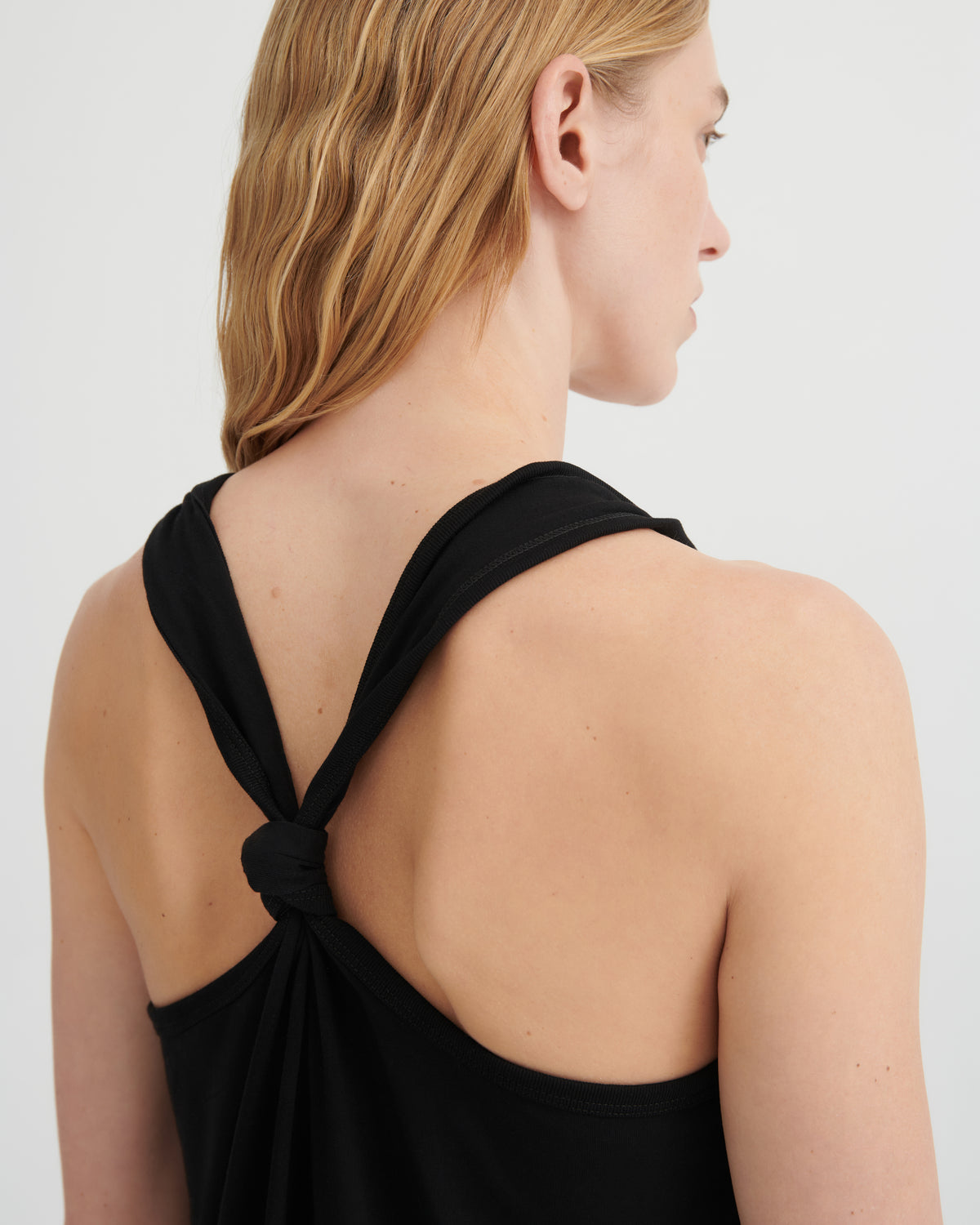 A woman shown from the back wearing a black sleeveless top with a knotted crossover back detail, against a plain light gray studio background.
