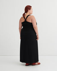 A woman with red hair tied back is shown from the back wearing a black halter-neck maxi dress and brown sandals against a plain white studio background.