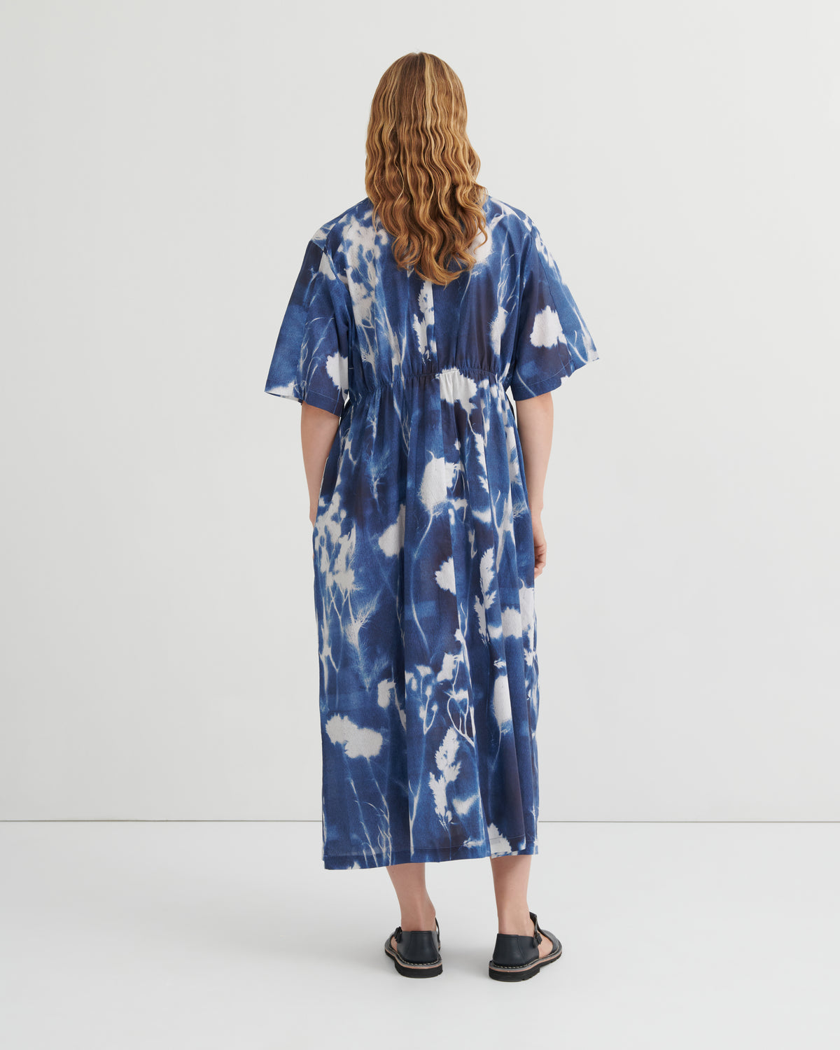 A woman viewed from behind wearing a loose, short-sleeve midi dress with a blue and white tie-dye pattern, paired with black flat sandals, against a plain white studio background.