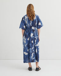 A woman viewed from behind wearing a loose, short-sleeve midi dress with a blue and white tie-dye pattern, paired with black flat sandals, against a plain white studio background.