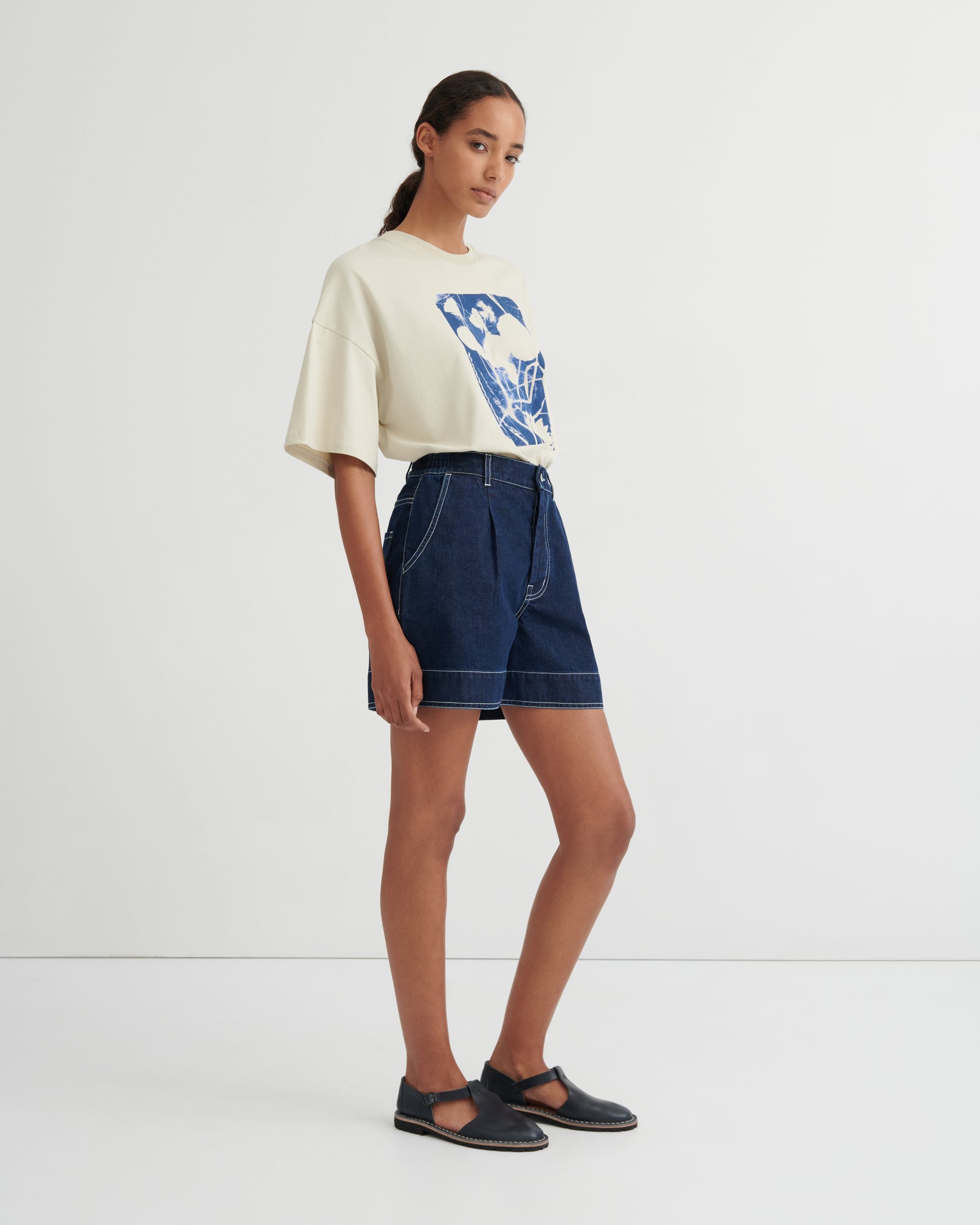 A model stands in a studio wearing a cream-colored oversized t-shirt with a blue graphic print, dark blue denim shorts, and dark navy Mary Jane flats.