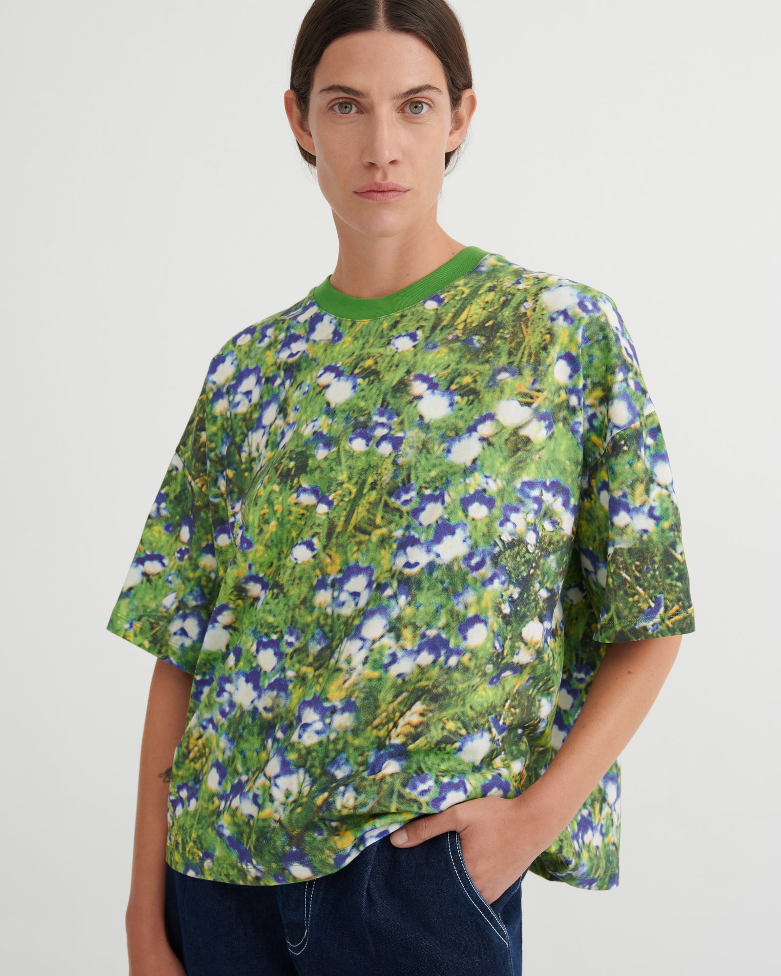 A model wearing a green short-sleeve crewneck t-shirt with a dense blue, white, and yellow floral print, paired with dark blue jeans, against a plain light gray background.