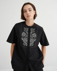 A model wearing a black short-sleeve button-up shirt with white embroidered floral details on the chest, photographed against a light gray studio background.