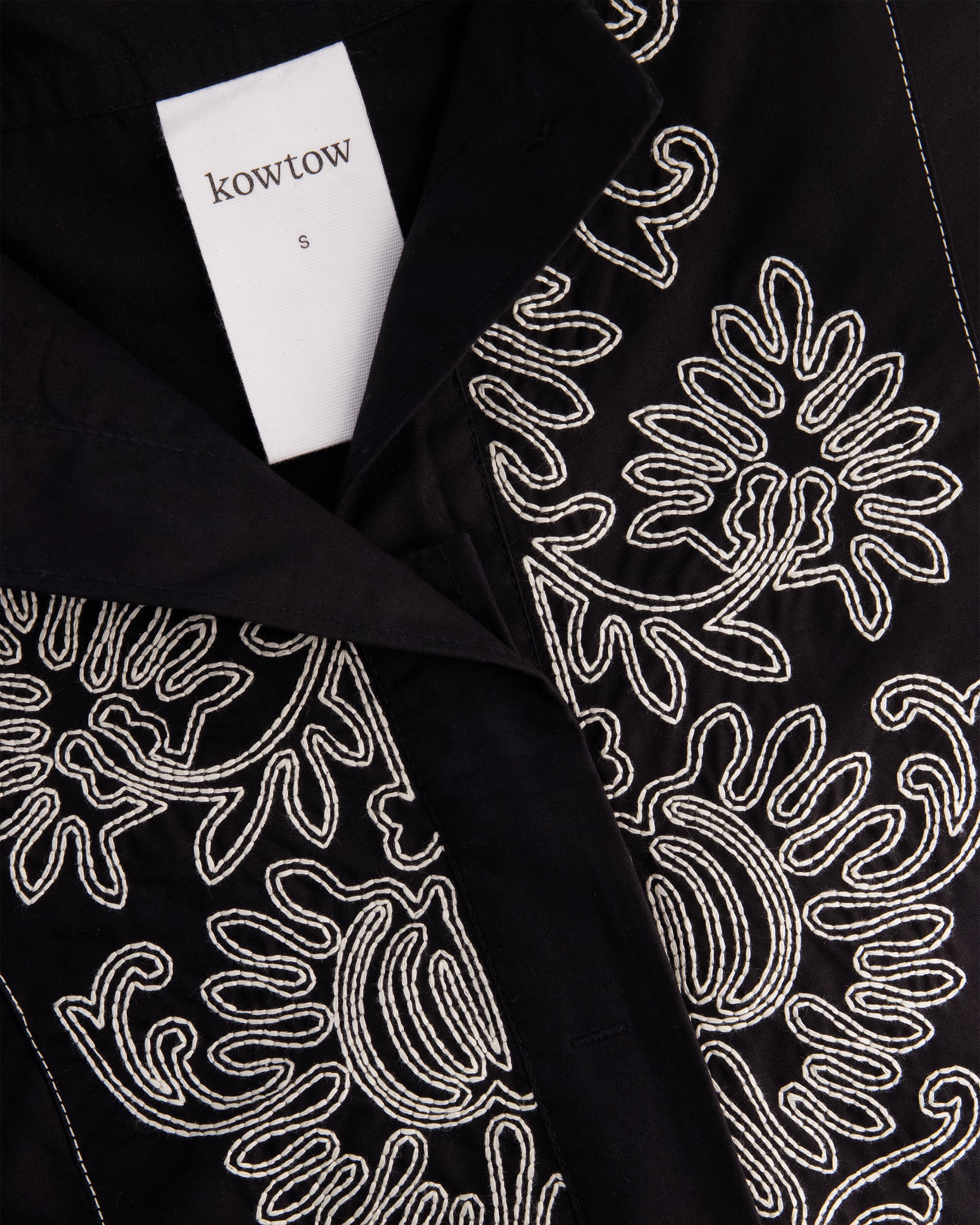 Close-up of a black garment with intricate white embroidered floral patterns, featuring a white brand label.