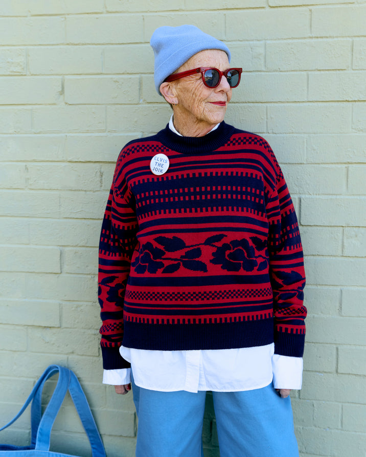 An elderly woman wearing a red and navy patterned knit sweater, a light blue beanie, red-framed sunglasses, and light blue pants, standing against a light beige brick wall.