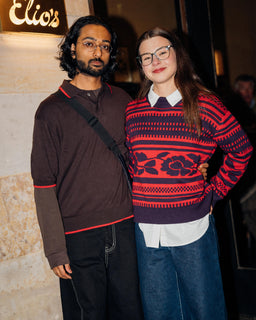 Two people standing together in front of a building entrance at night. The person on the right wears a red and navy patterned sweater over a white collared shirt and blue jeans, while the person on the left wears a dark brown long-sleeve polo with red trim and black pants.