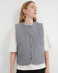 A woman wearing a sleeveless quilted vest with vertical blue, beige, and white stripes over a white short-sleeve top, photographed against a plain light gray wall.
