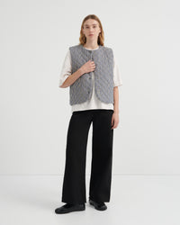 A model stands against a plain white studio background wearing a sleeveless textured gray vest over a white short-sleeve top, black wide-leg trousers, and black shoes.