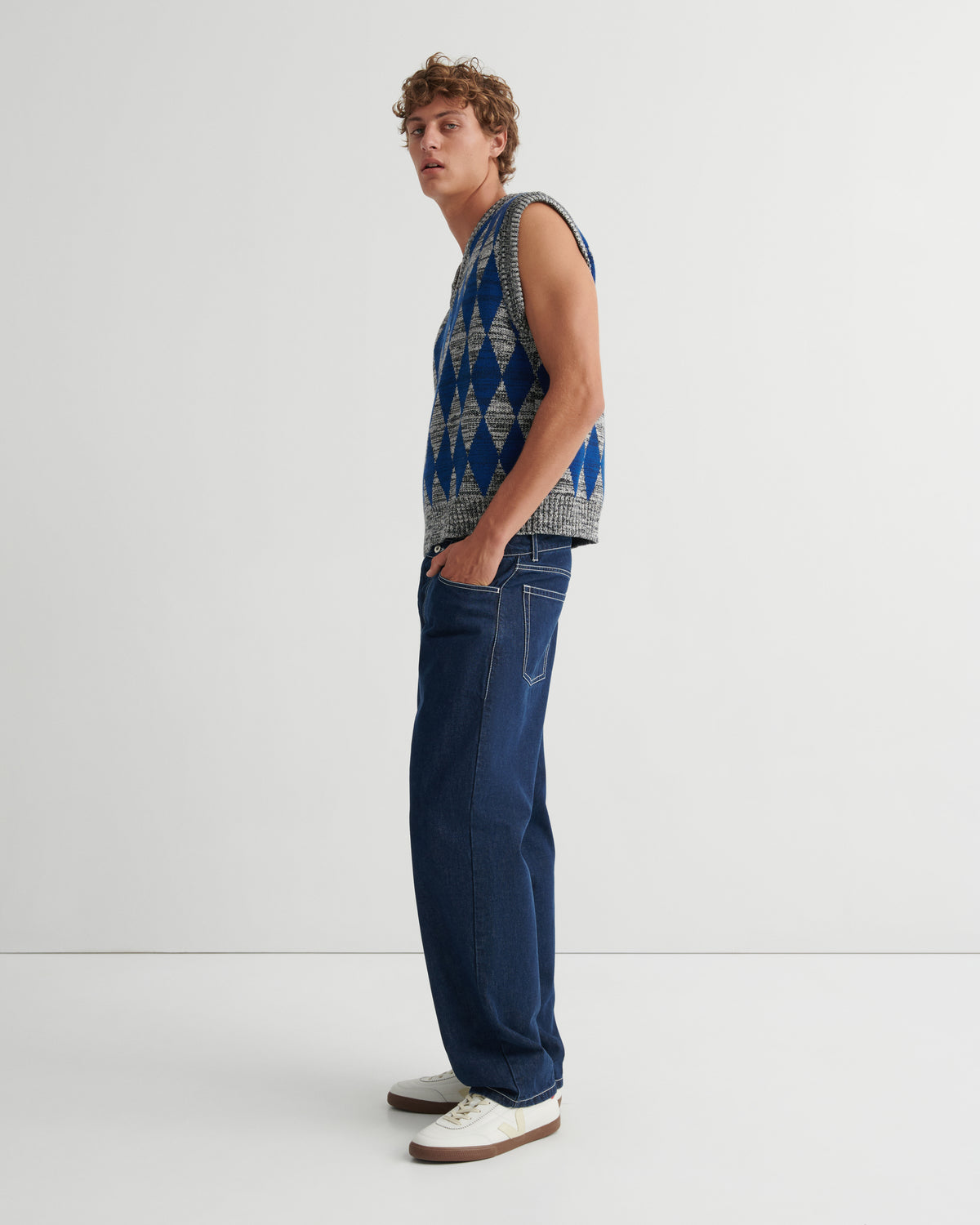 A man stands in profile wearing a sleeveless knit vest with a blue, gray, and white geometric pattern, dark blue jeans, and white sneakers with brown soles.