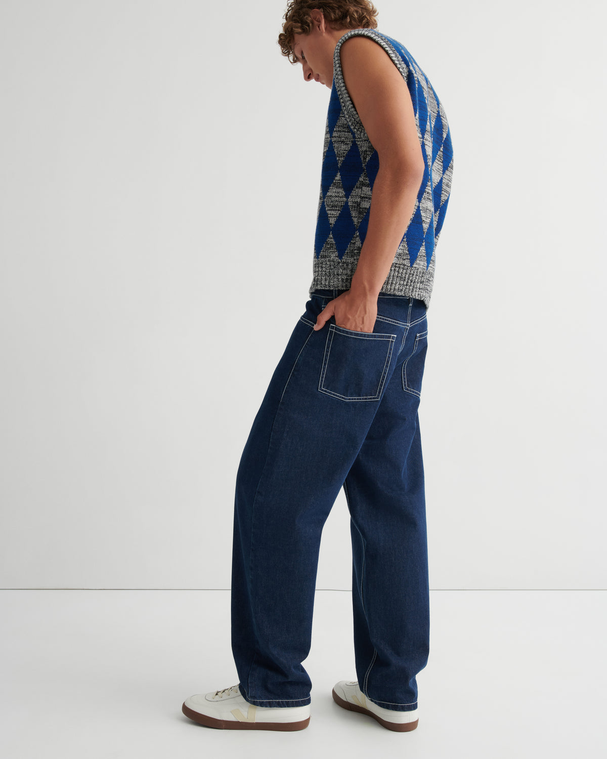 A model shown from the side wearing a blue and white patterned sleeveless knit vest, dark blue wide-leg jeans, and white sneakers.