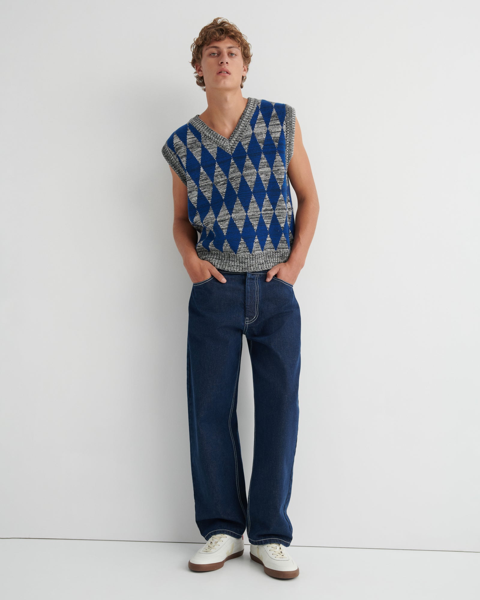 A model wearing a sleeveless knit sweater vest with a blue and gray geometric diamond pattern, dark blue jeans, and white sneakers.