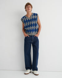 A model wearing a sleeveless knit sweater vest with a blue and gray geometric diamond pattern, dark blue jeans, and white sneakers.