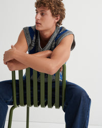 A young man with curly brown hair sits on a green slatted chair, wearing a sleeveless knit vest with a blue and gray pattern over a light blue shirt and dark blue jeans.