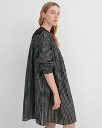A woman modeling an oversized black and white checkered dress with solid black shoulder yokes, photographed against a neutral light gray studio background.