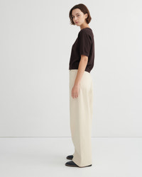 A woman stands in profile wearing a dark brown short-sleeve top, cream wide-leg trousers, and dark loafers against a clean white studio background.