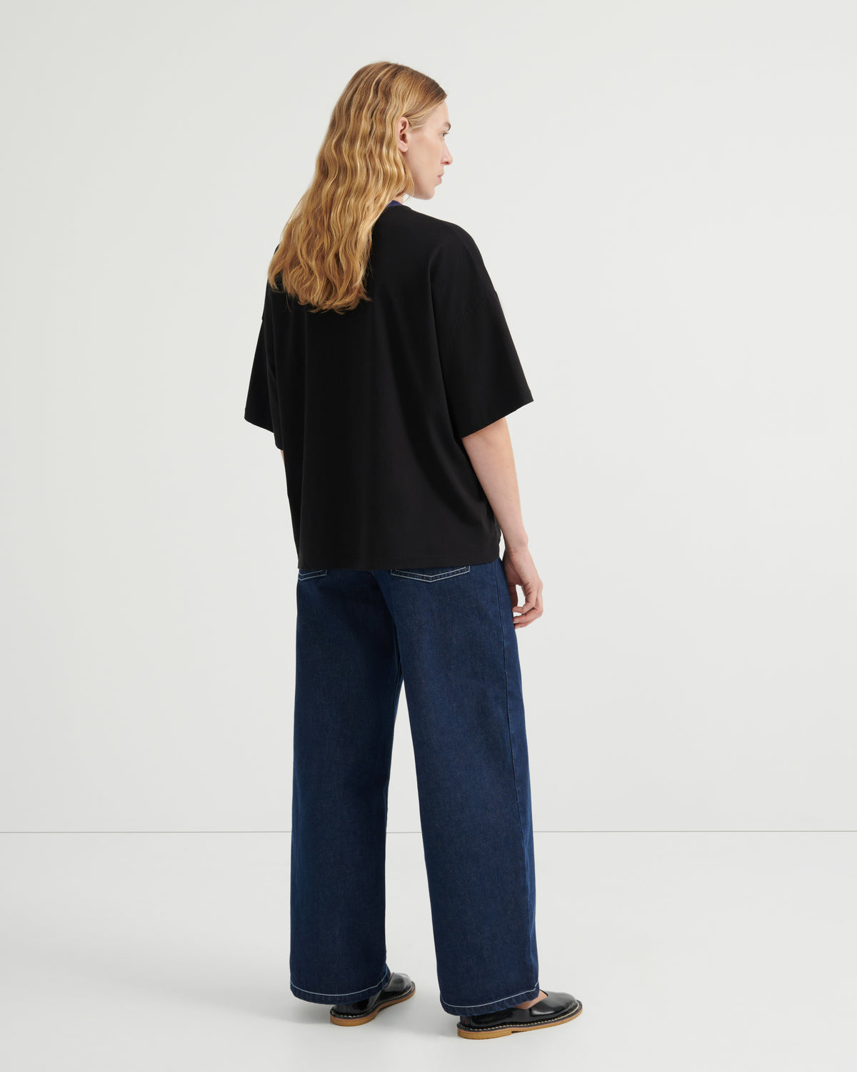 A woman with long blonde hair is shown from the back wearing an oversized black t-shirt and wide-leg dark blue jeans, standing against a plain light gray studio background.