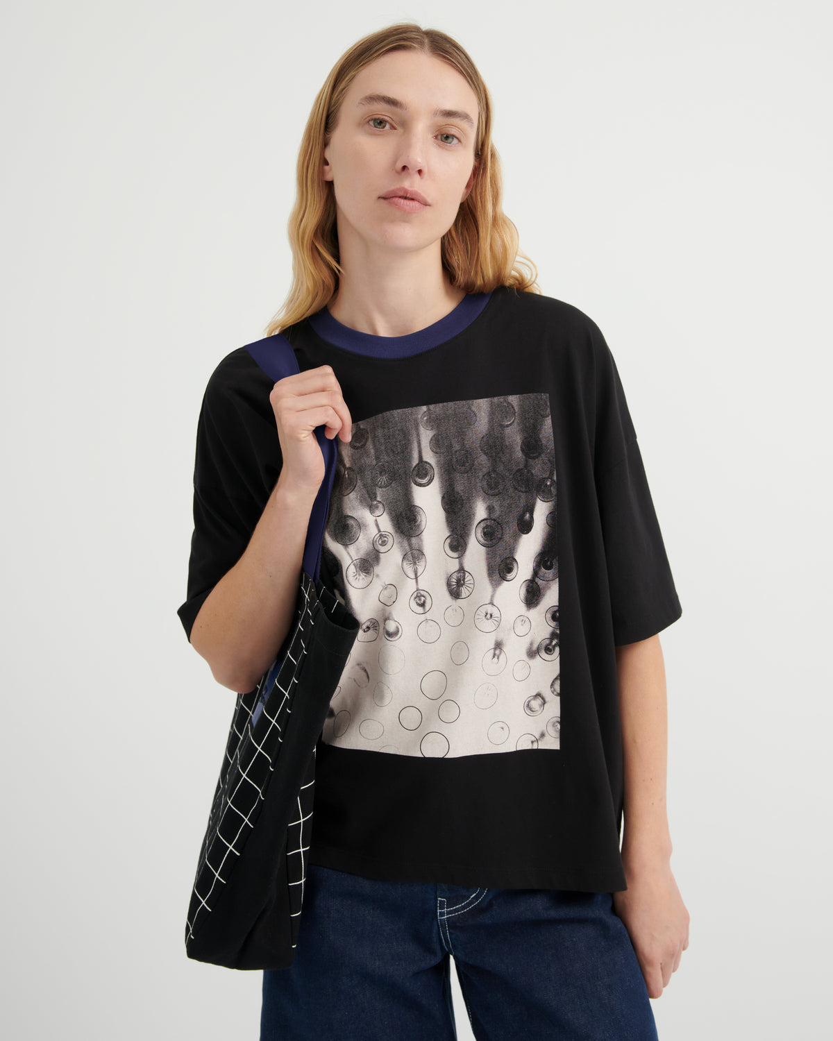 A woman modeling a black oversized graphic t-shirt with a dark blue ribbed collar, paired with dark blue jeans and carrying a black tote bag with white grid lines against a light gray studio background.