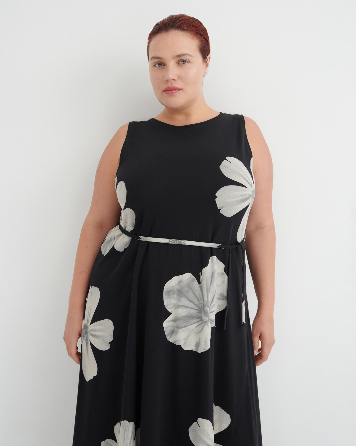 A woman modeling a sleeveless black midi dress with a silver waist tie and large white floral print, set against a plain light gray background.