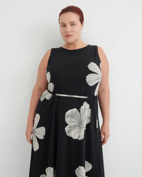 A woman modeling a sleeveless black midi dress with a silver waist tie and large white floral print, set against a plain light gray background.