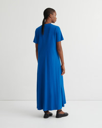A woman wearing a bright blue short-sleeve maxi dress, shown from the back in a studio setting with a white background.
