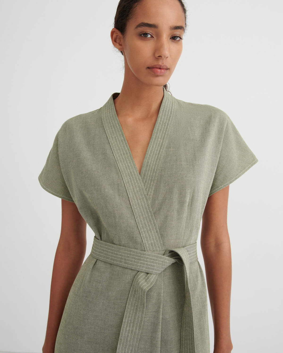 A woman wearing a sage green short-sleeve wrap dress with a matching tie belt, photographed against a plain light gray studio background.