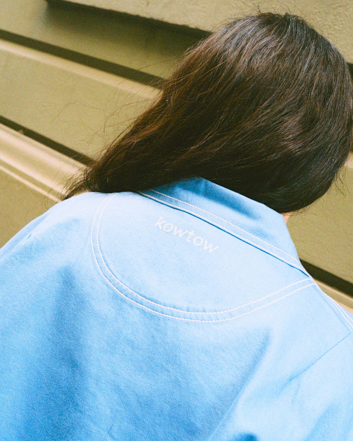 A person viewed from behind wearing a light blue Kowtow jacket with their dark brown hair visible against a beige wall background.