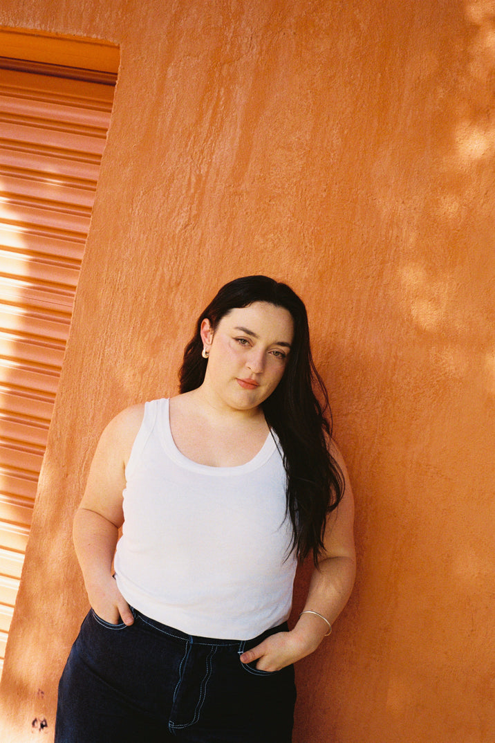 A woman with long dark hair stands against a textured orange wall, wearing a white tank top and dark blue jeans.