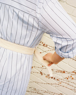 A close-up lifestyle shot of a person wearing a light blue and white pinstriped shirt with a cream-colored waist belt.