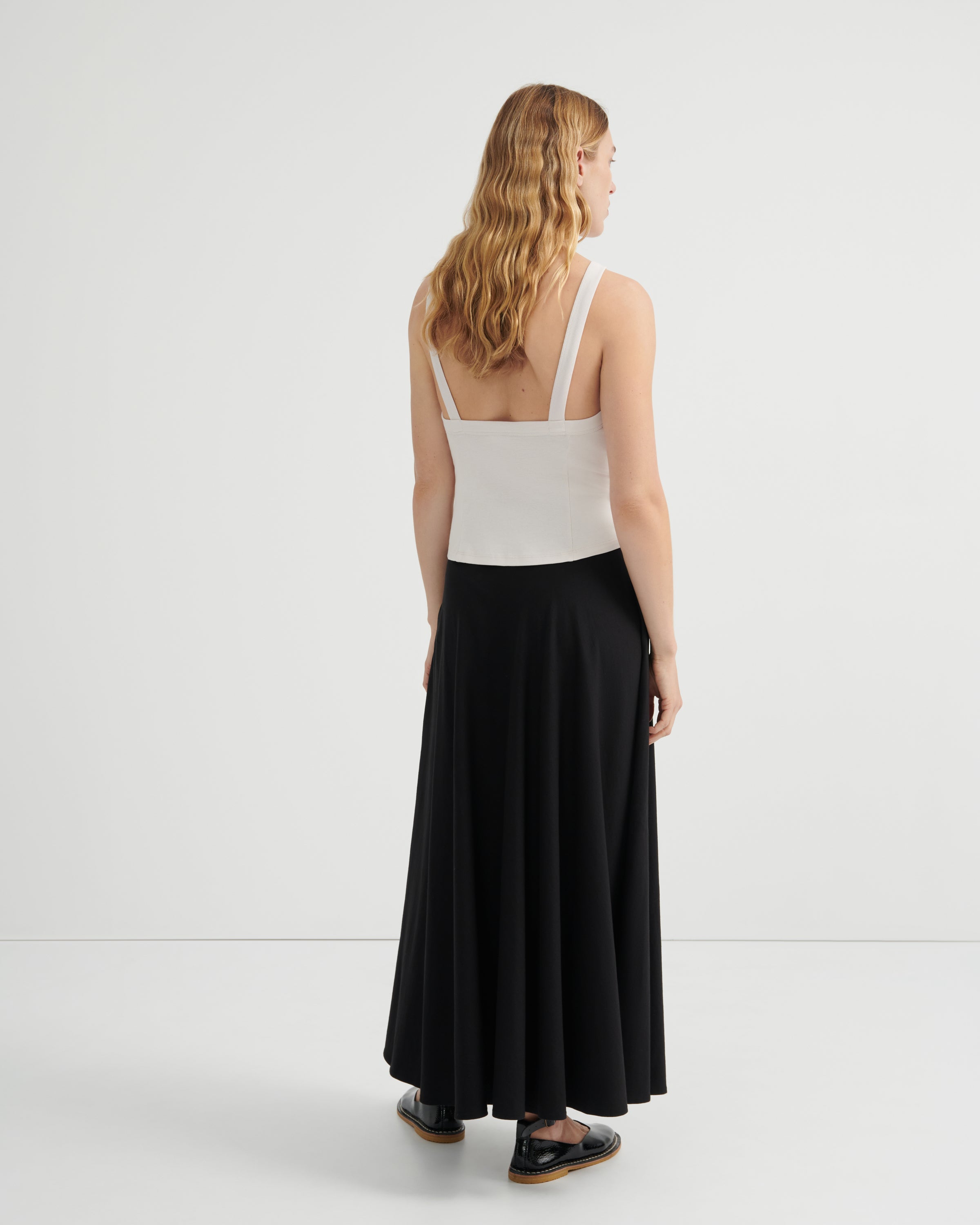 A woman is shown from the back wearing a white sleeveless top with wide straps and a long black skirt, standing against a plain light gray studio background.