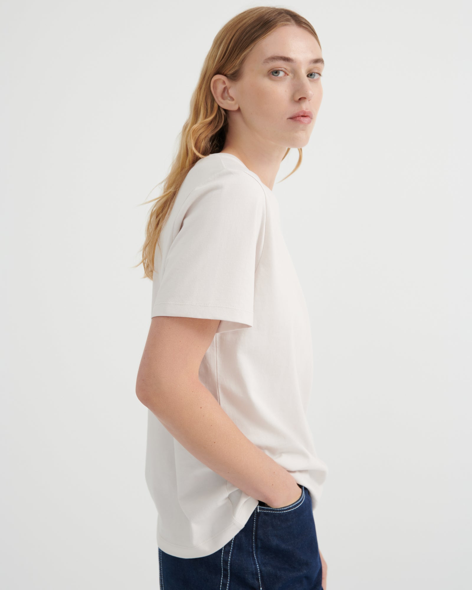 A woman with blonde hair is shown in profile wearing a plain white short-sleeve crewneck t-shirt and dark blue jeans against a light gray studio background.
