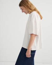 A woman wearing an oversized off-white short-sleeve t-shirt and dark blue jeans, shown from a side profile against a clean white studio background.
