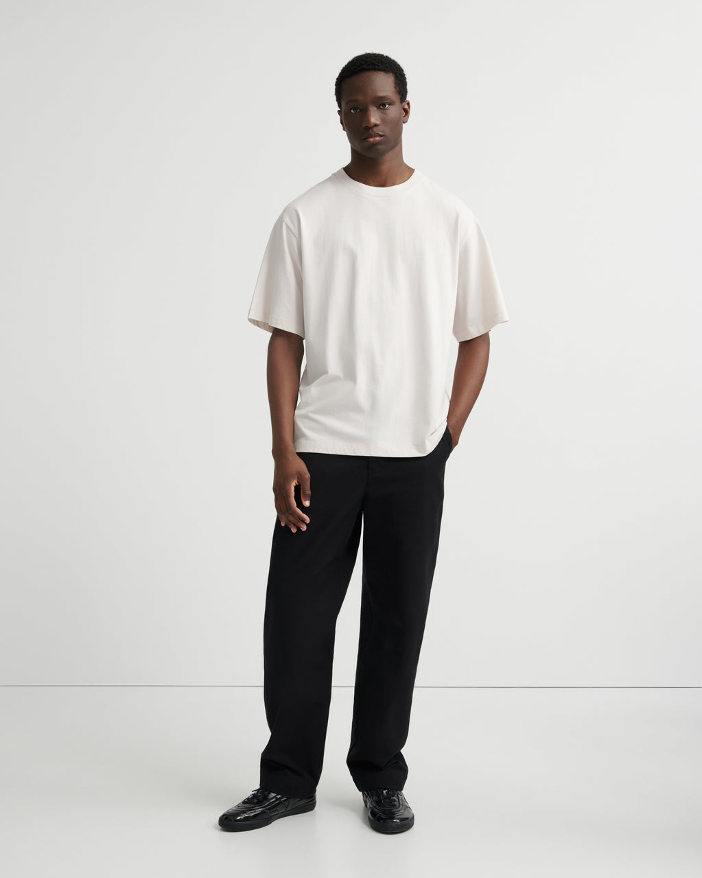A man modeling a loose-fitting off-white short-sleeve t-shirt and black trousers against a clean white studio background.