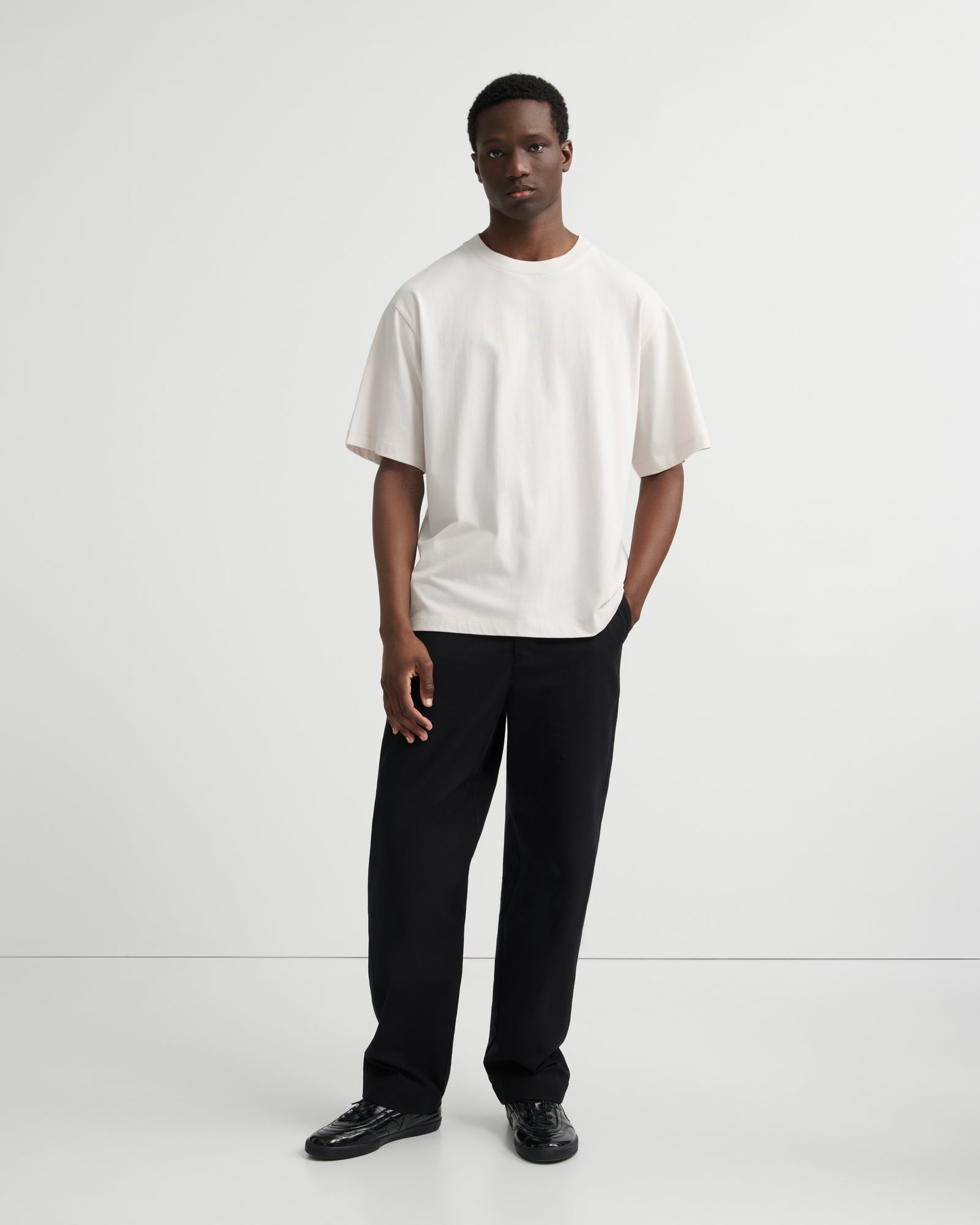 A man modeling a loose-fitting off-white short-sleeve t-shirt and black trousers against a clean white studio background.