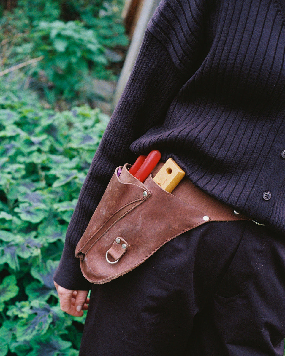 A brown leather waist tool pouch worn on a person's hip, holding red-handled tools, shown outdoors against a blurred green garden background.