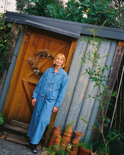 A woman with short blonde hair wearing a blue denim jumpsuit stands in front of a rustic wooden door of a corrugated metal shed, surrounded by green garden foliage and potted plants.