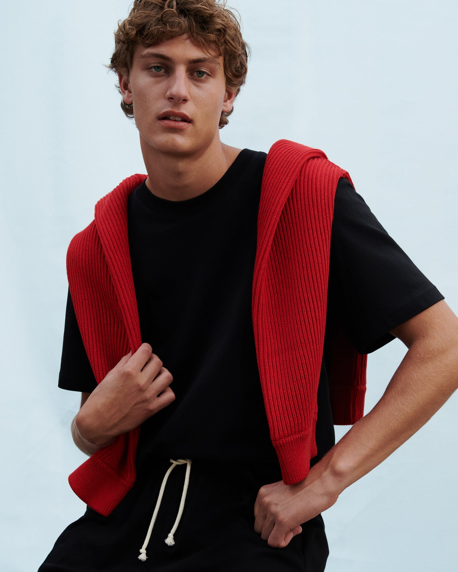 Model standing against a blue background wearing a relaxed black t-shirt with a red sweater draped over his shoulders.