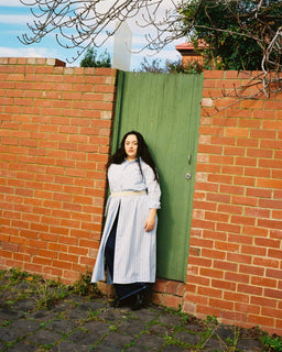 A woman stands in front of a red brick wall and a green wooden door, wearing a long white and blue striped dress with a beige belt. Bare tree branches hang from above, and the ground is paved with cobblestones.