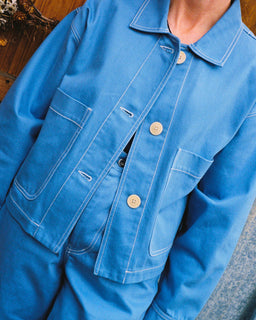 A close-up of a person wearing a bright blue work jacket with white contrast stitching and light-colored buttons, paired with matching blue pants.