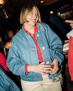 A woman smiling in a social setting, wearing a light blue denim jacket over a red collared shirt and beige pants, holding a glass.
