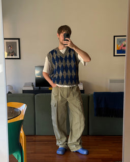 A man takes a mirror selfie in a living room, wearing a navy and beige argyle sweater vest over a white t-shirt, loose khaki cargo pants, and blue slip-on shoes.