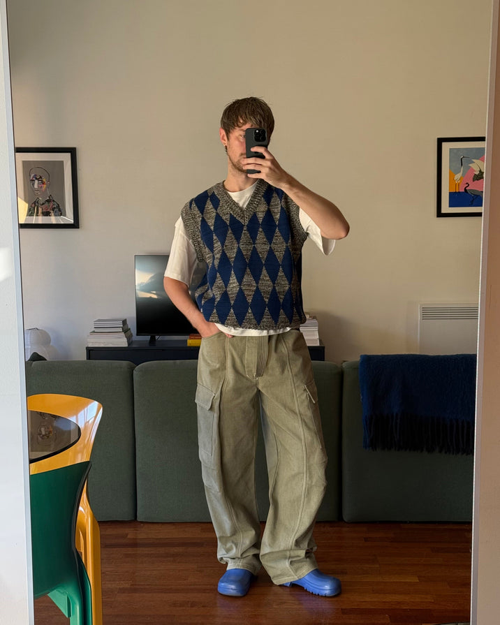 A man takes a mirror selfie in a living room, wearing a navy and beige argyle sweater vest over a white t-shirt, loose khaki cargo pants, and blue slip-on shoes.
