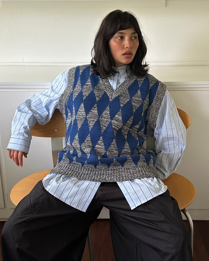 A model sits on a wooden chair wearing a blue and gray argyle knit sweater vest over a light blue and white striped button-down shirt, paired with wide-leg black trousers.