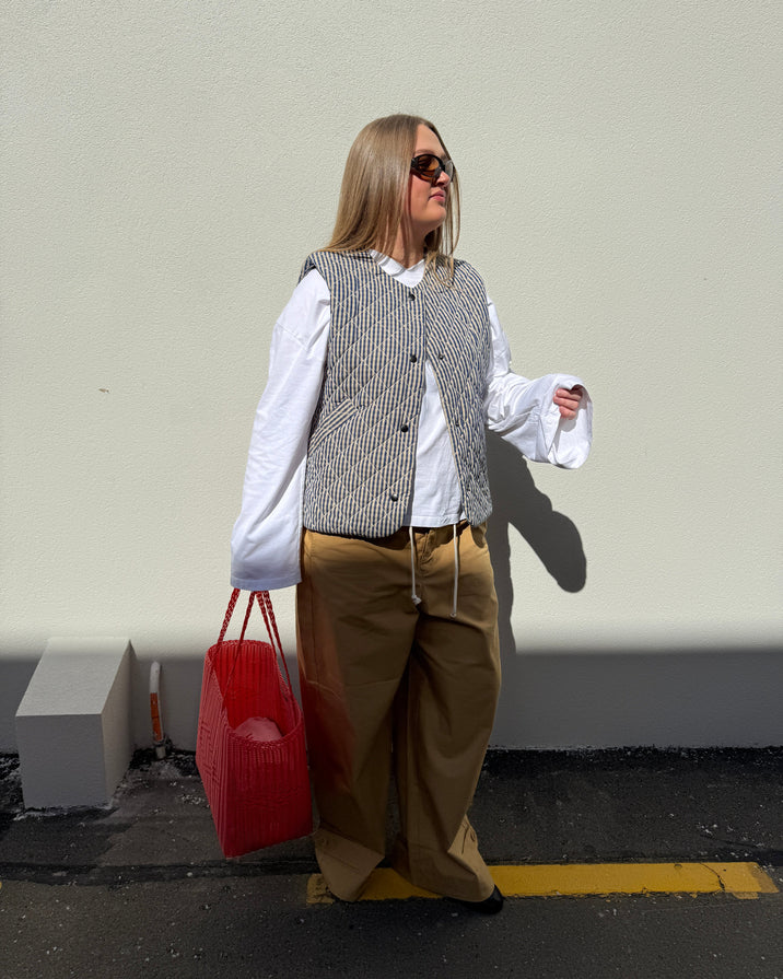 A woman stands outdoors against a plain light wall wearing a gray and white patterned vest over a white long-sleeve blouse, tan wide-leg pants, and black sunglasses. She is holding a bright red tote bag.