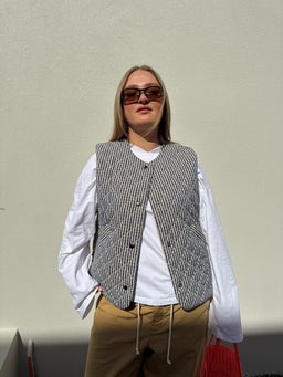 A woman wearing a gray and white striped quilted vest over a white long-sleeve shirt, tan pants, and dark sunglasses, standing against a plain light beige wall.