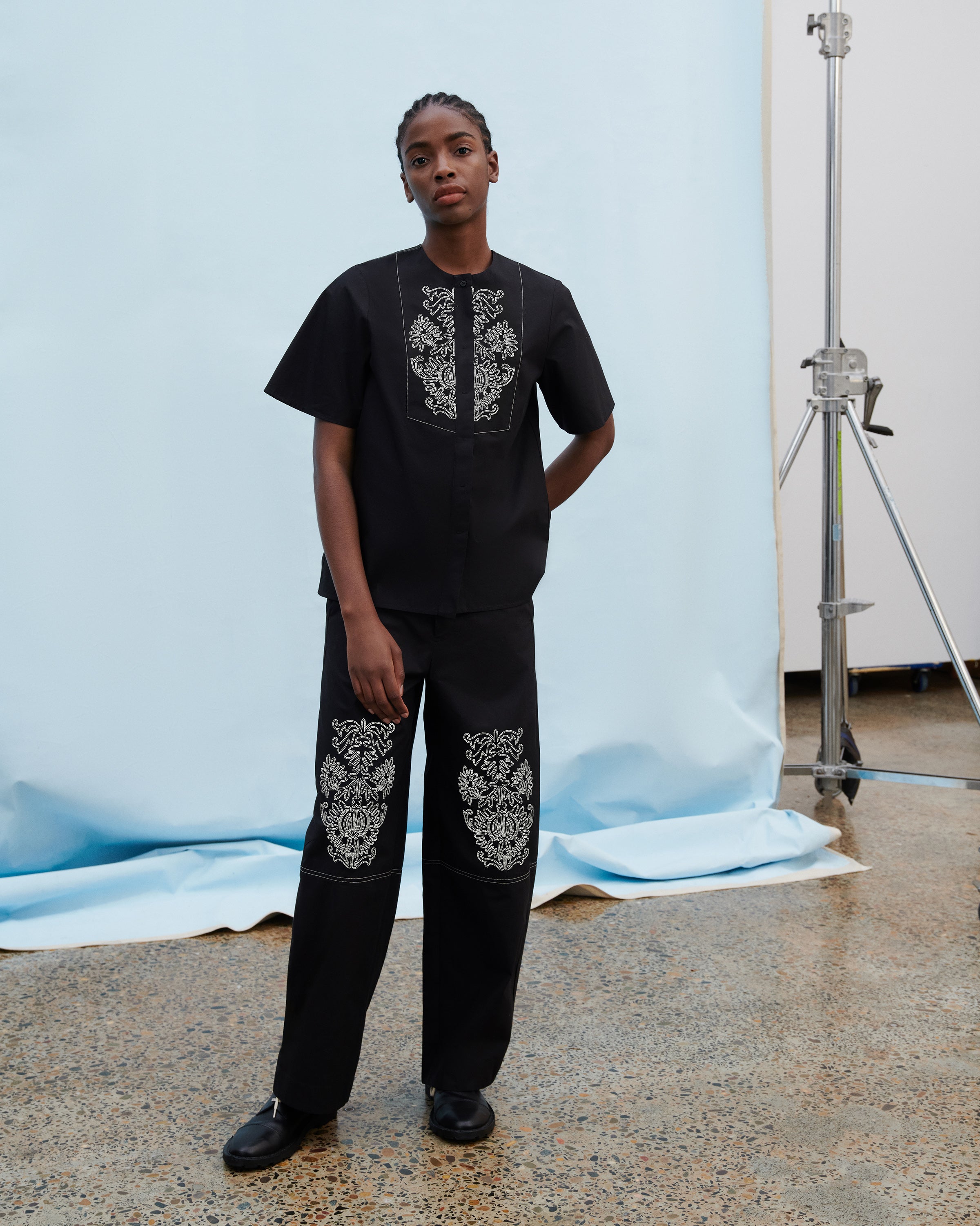 A model stands in a studio wearing a black short-sleeve tunic and matching wide-leg pants, both featuring ornate white embroidered patterns on the chest and upper legs. The background shows a light blue fabric backdrop with a studio light stand visible on the right.
