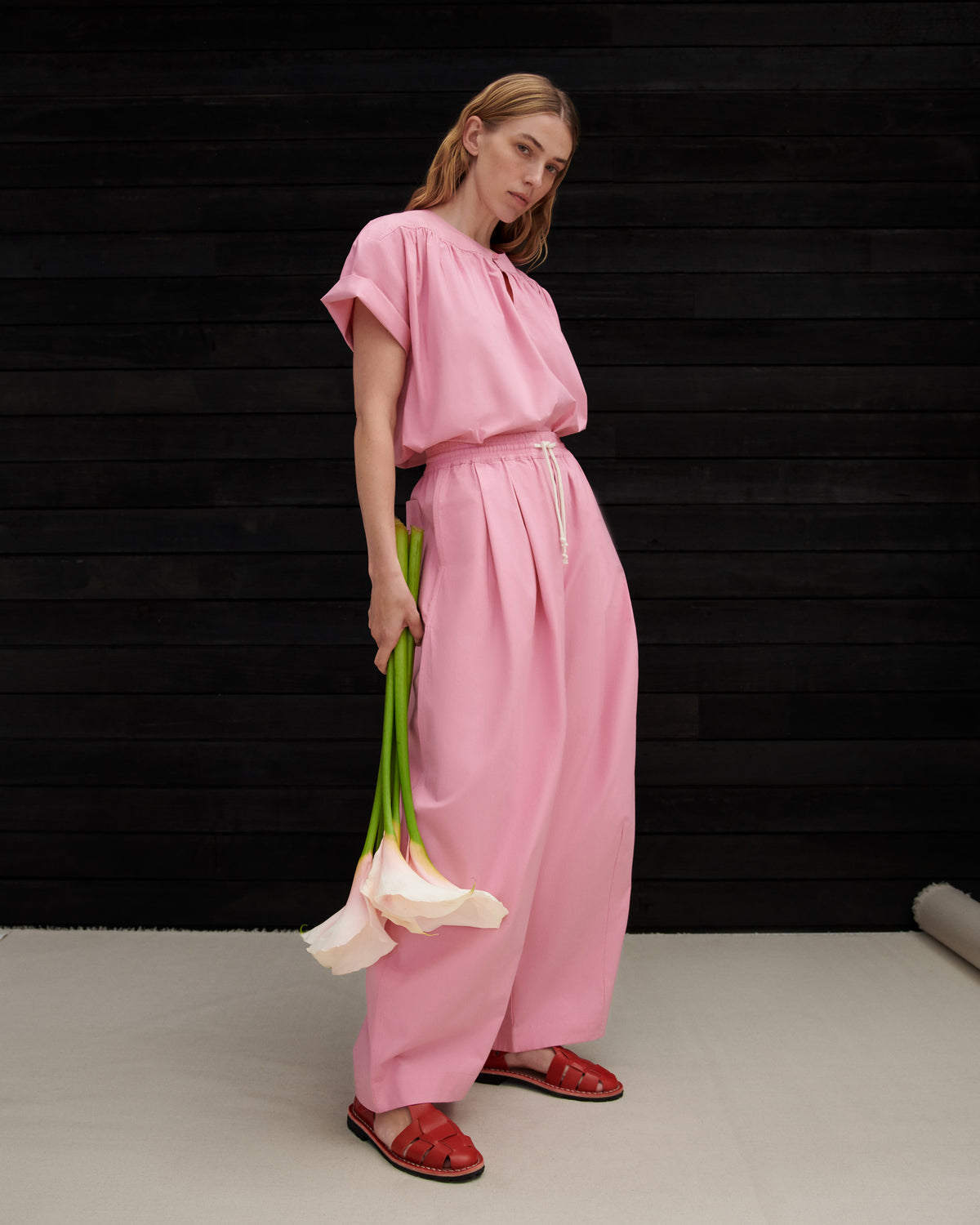A woman modeling a light pink two-piece linen set consisting of a short-sleeve top and matching wide-leg trousers, holding a bunch of white calla lilies against a dark wood-paneled background.