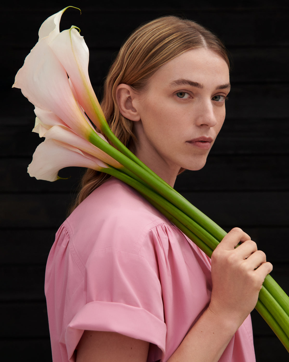 A woman with blonde hair wearing a pink top holds a large bouquet of pale pink and white calla lilies against a dark wooden background.