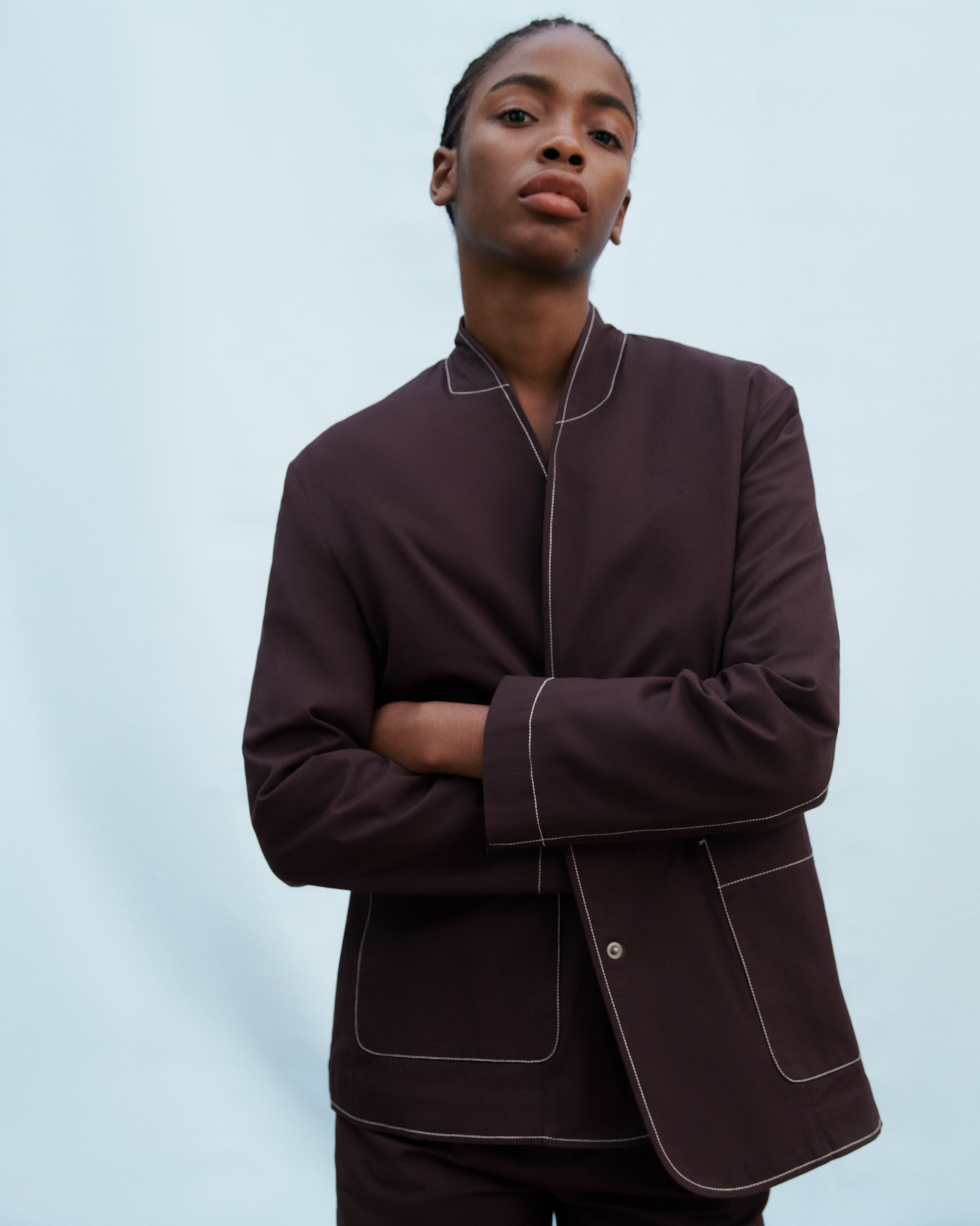 A model wearing a dark  brown colored blazer with contrasting light topstitching posed against a solid light blue background.