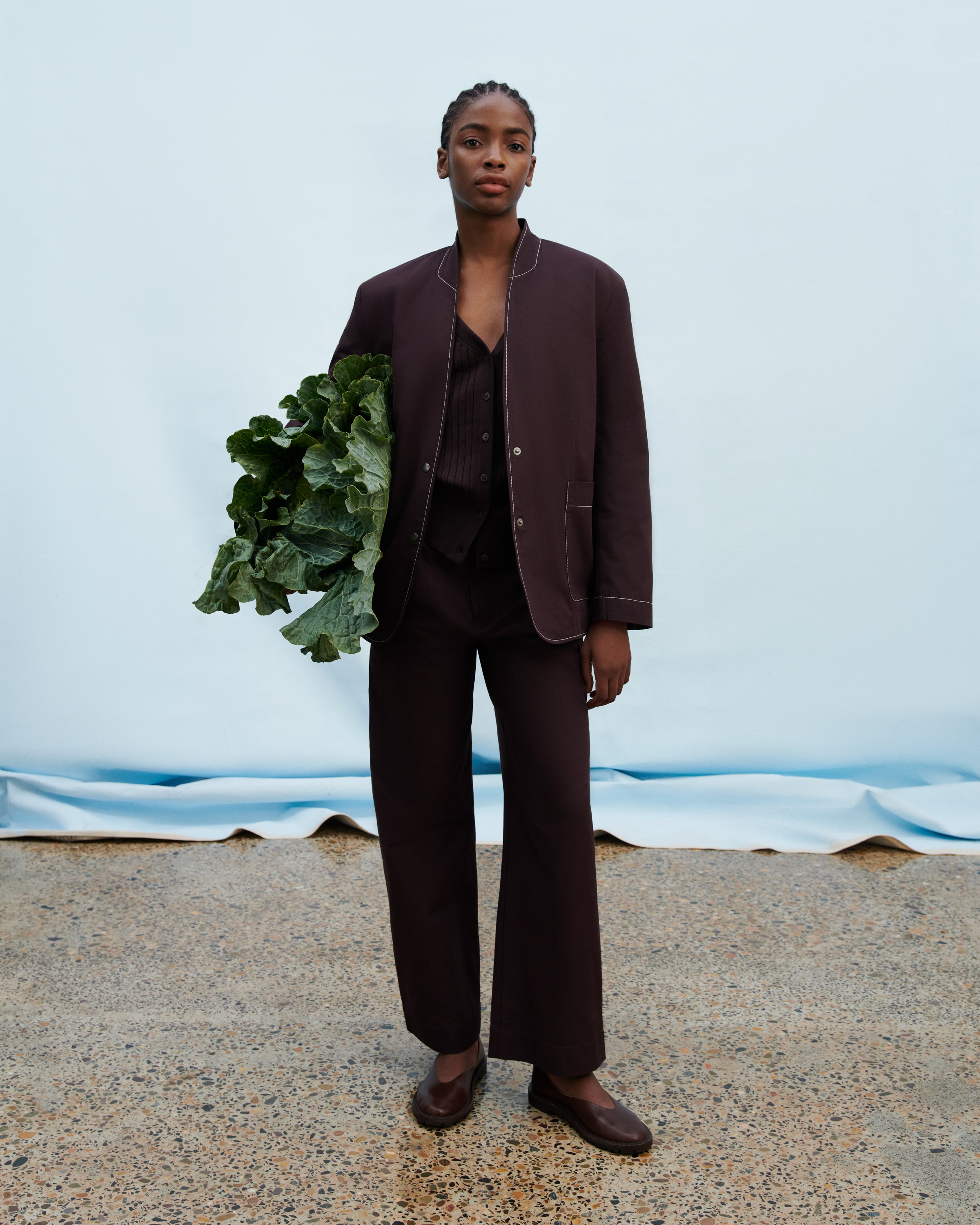 A model stands against a light blue backdrop wearing a dark brown tailored suit with a blazer, vest, and wide-leg trousers, holding a bunch of green leafy vegetables.